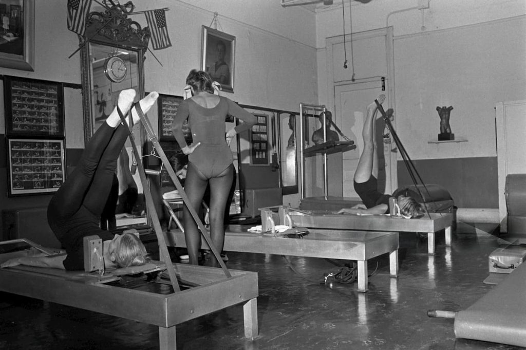 Tri devojke na pilates reformeru rade vežbe, slika je crno bela sa početka 20.veka.